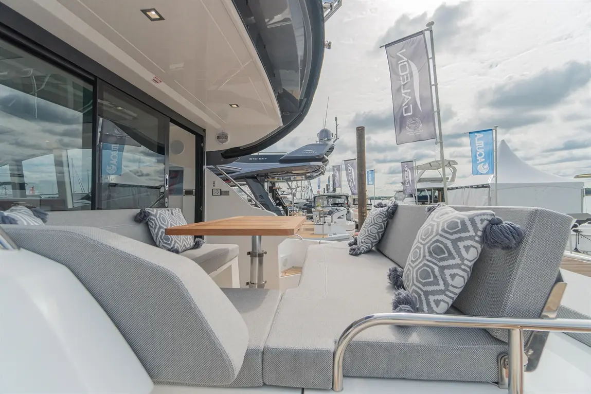 Stylish yacht lounge area with grey cushions and wooden table, showcasing nearby boats and flags under a partly cloudy sky.